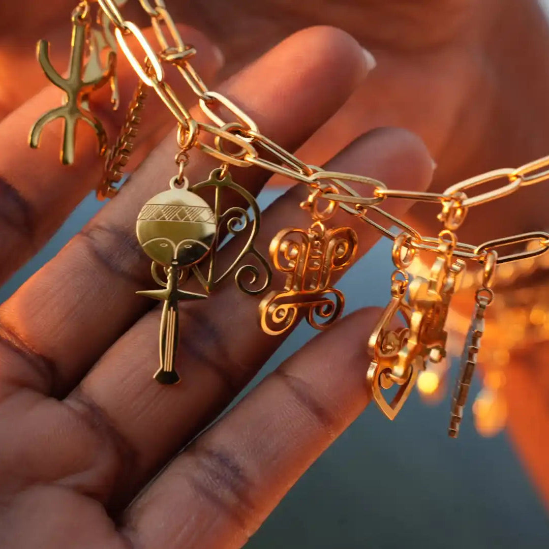 Gold African charm bracelet with Adinkra symbols and Baule fertility doll, handcrafted chain link design, held in hand against blurred background. Cultural jewelry, Afro-inspired accessory, meaningful storytelling bracelet