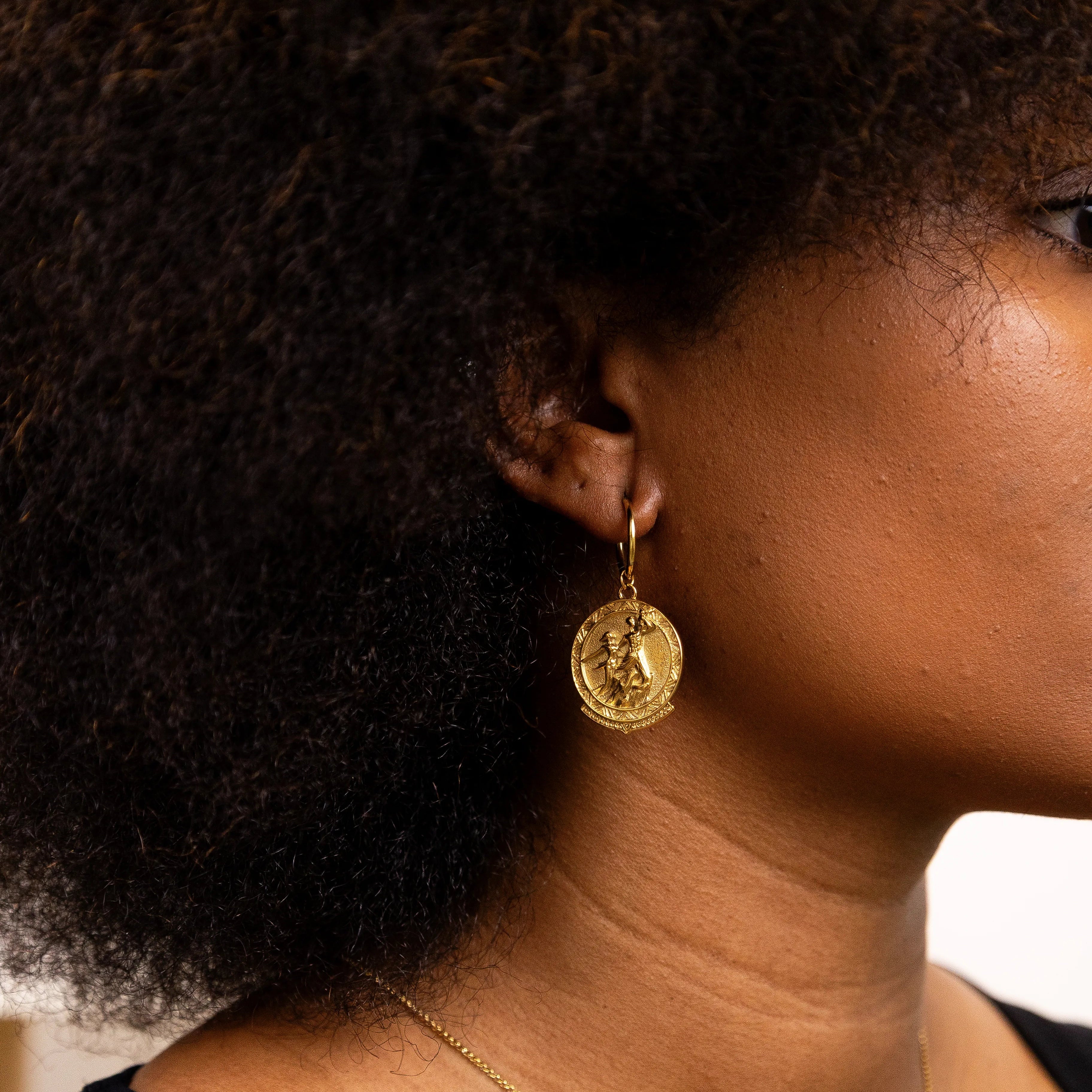 Close-up of a person wearing gold earrings and a necklace with a blurred background. coin earrings with the Senegal renaissance monument design.
