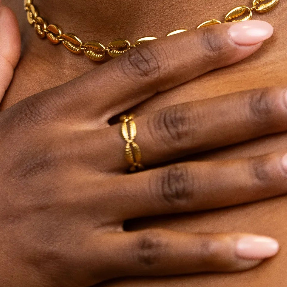 Close-up of a person wearing a gold cowries necklace and ring, with a neutral background.