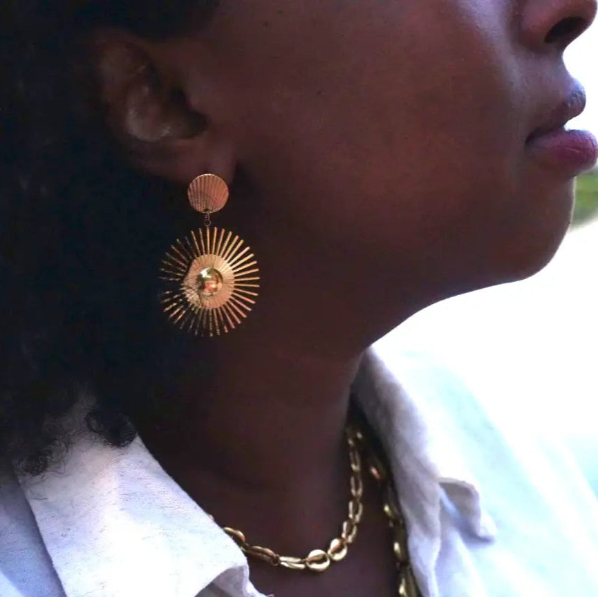 Close-up of a person wearing gold earrings and a necklace with a blurred background - Tilo La Earrings - sun earrings doore dasi